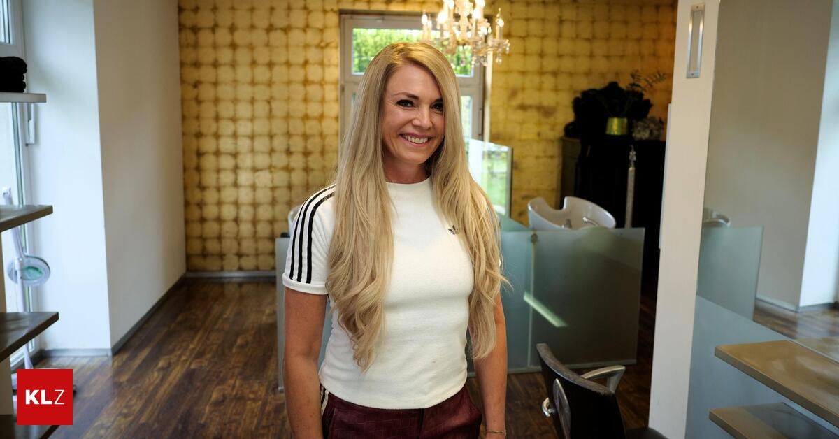 A woman with blonde hair stands in a salon, smiling. She wears a white shirt with black stripes. Behind her, a glass partition and a chandelier are visible.