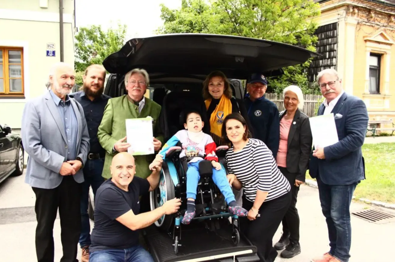 A group of adults and a child in a wheelchair are standing next to a car with its trunk open. The group is smiling and posing for a photo.