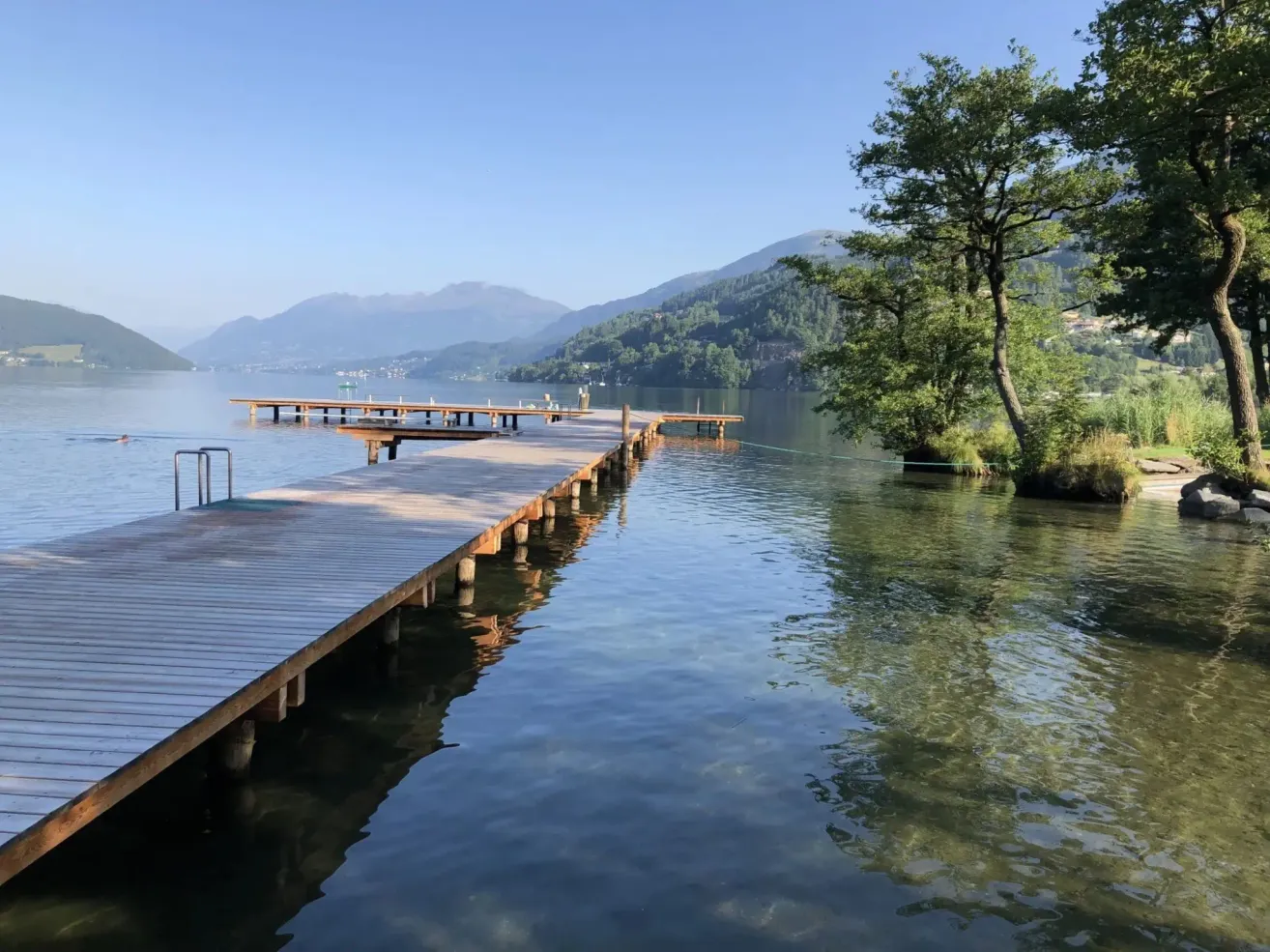 Ein ruhiger See mit einem hölzernen Steg, der sich ins Wasser erstreckt, einem Baum rechts und Bergen in der Ferne unter einem klaren blauen Himmel.