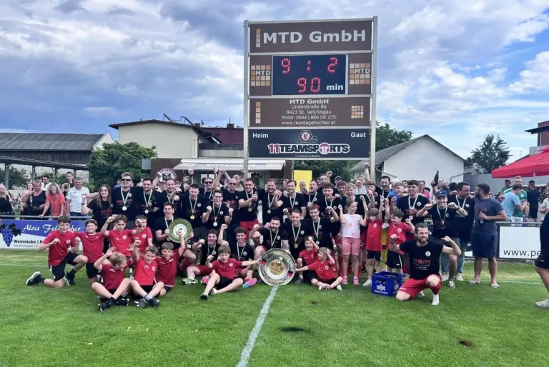 Eine Gruppe junger Fußballspieler in roten Trikots und mit Medaillen posiert für ein Foto auf dem Feld. Ein Pokal und ein Schild mit MTD GmbH stehen vor ihnen.
