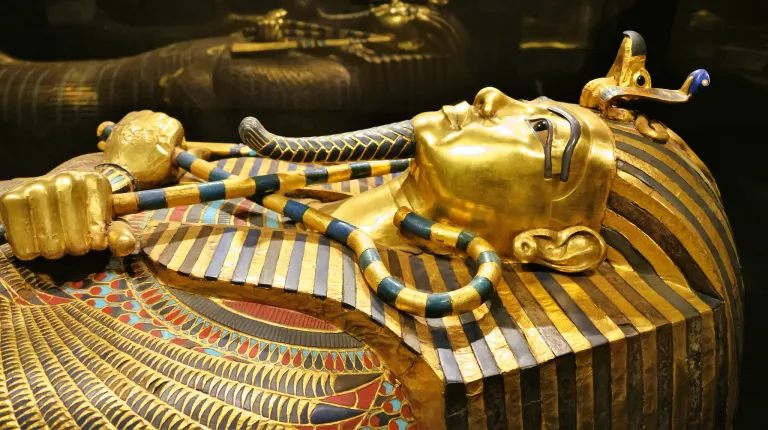 Ein goldener Sarkophag liegt auf einer Plattform, verziert mit komplizierten Mustern. Ein goldener Maske bedeckt das Gesicht, und ein schlangenartiger Kopfschmuck ist auf dem Kopf.