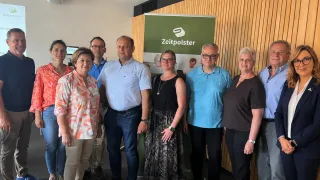 Eight people are posing for a photo in front of a wooden wall with a green banner that reads Zeitpolster. They all appear to be smiling and looking forward.
