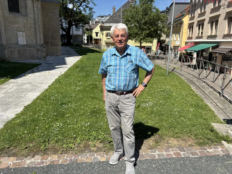Ein älterer Mann steht auf einer Kopfsteinpflasterstraße, lächelt und schaut nach rechts. Hinter ihm ist ein üppiger Rasen und ein gelbes Gebäude zu sehen.