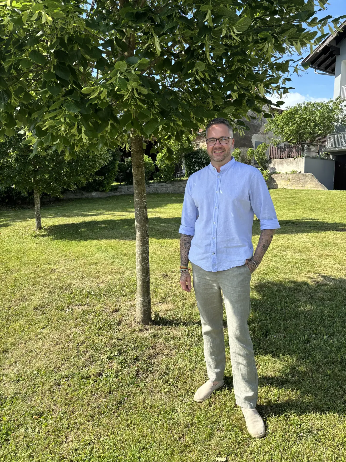 Ein Mann mit Tätowierungen steht lächelnd im Garten, mit den Händen in den Taschen. Hinter ihm steht ein kleiner Baum und ein Haus.