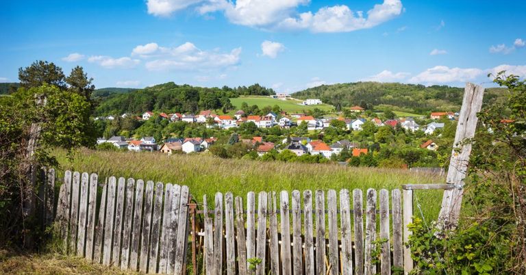 Ein Dorf in einem Tal, umgeben von üppiger Grünfläche und Häusern mit roten Dächern, unter einem blauen Himmel mit verstreuten Wolken.