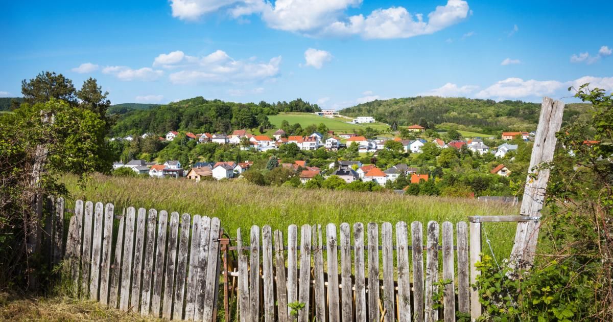 A town nestled in a valley, surrounded by lush greenery and houses with red roofs, under a blue sky with scattered clouds.