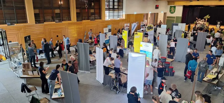 Eine Indoor-Veranstaltung mit Menschen an Ständen und Tischen, Bannern und Leuten, die herumlaufen. Holzwände und Fenster.