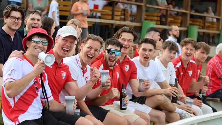 Several men wearing red and white jerseys are cheering at a sports event, holding cups. They are seated in a stadium with a wooden bench behind them and some spectators in the background.