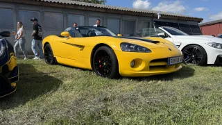 A bright yellow sports car is parked in front of a building with solar panels. A man stands nearby, while others look out of the building.