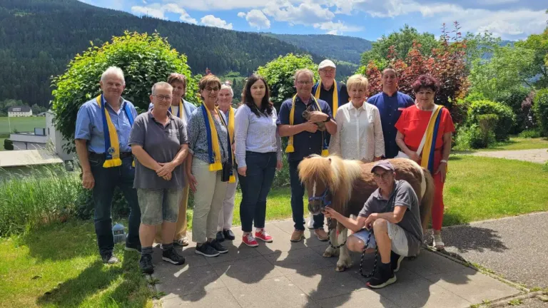 Eine Gruppe von Menschen steht draußen in der Nähe eines kleinen Pferdes, gekleidet in Freizeitkleidung, einige tragen Schals. Dahinter befinden sich Bäume und Berge.