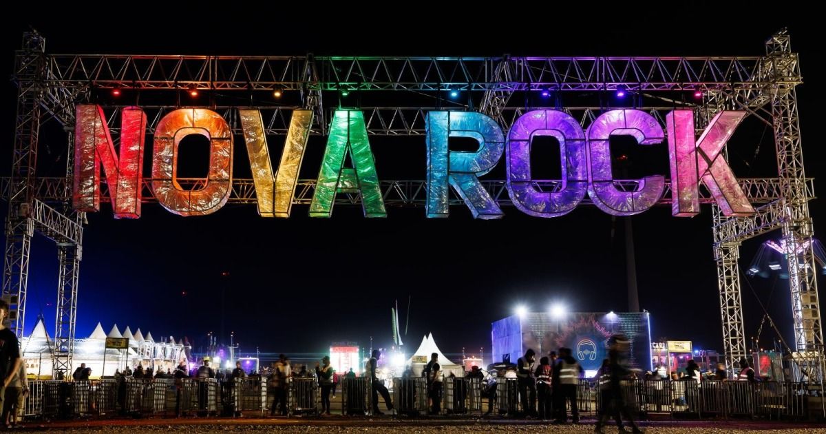 Der Nova Rock Festival-Eingang bei Nacht, mit bunt beleuchteten Buchstaben. Eine Menschenmenge versammelt sich vor dem Eingang, mit Zelten und Lichtern im Hintergrund.