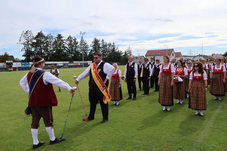 A group of people dressed in traditional costumes are standing on a grass field. They seem to be performing for an event. A man is holding a long rope and is walking beside a man in a hat. Behind them are trees, a house, and a street light.