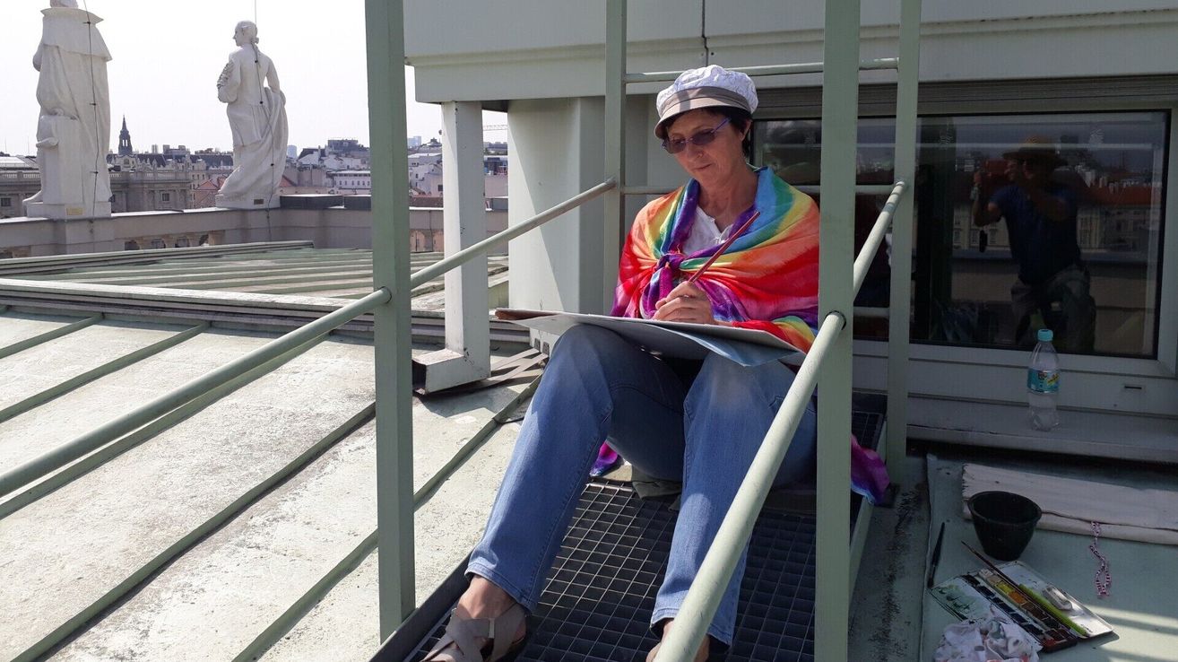 An artist is sitting on a rooftop, sketching on a paper. She is wearing a rainbow scarf and holding a pencil. The rooftop has metal railings and a metal grate. Behind her, there is a white statue.