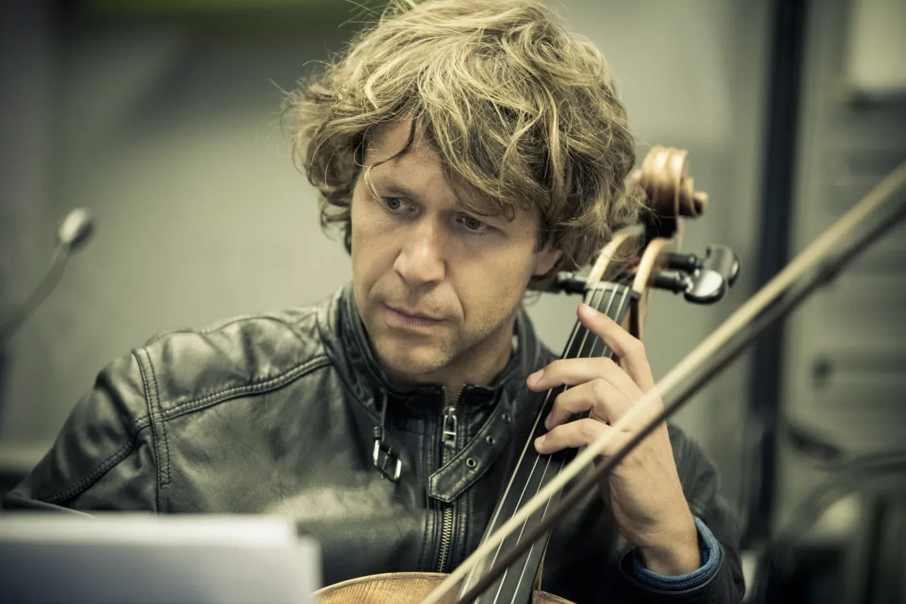A man with curly blonde hair, wearing a black leather jacket, is playing the cello. He is looking down and holding the instrument with both hands.