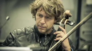 A man with curly blonde hair, wearing a black leather jacket, is playing the cello. He is looking down and holding the instrument with both hands.