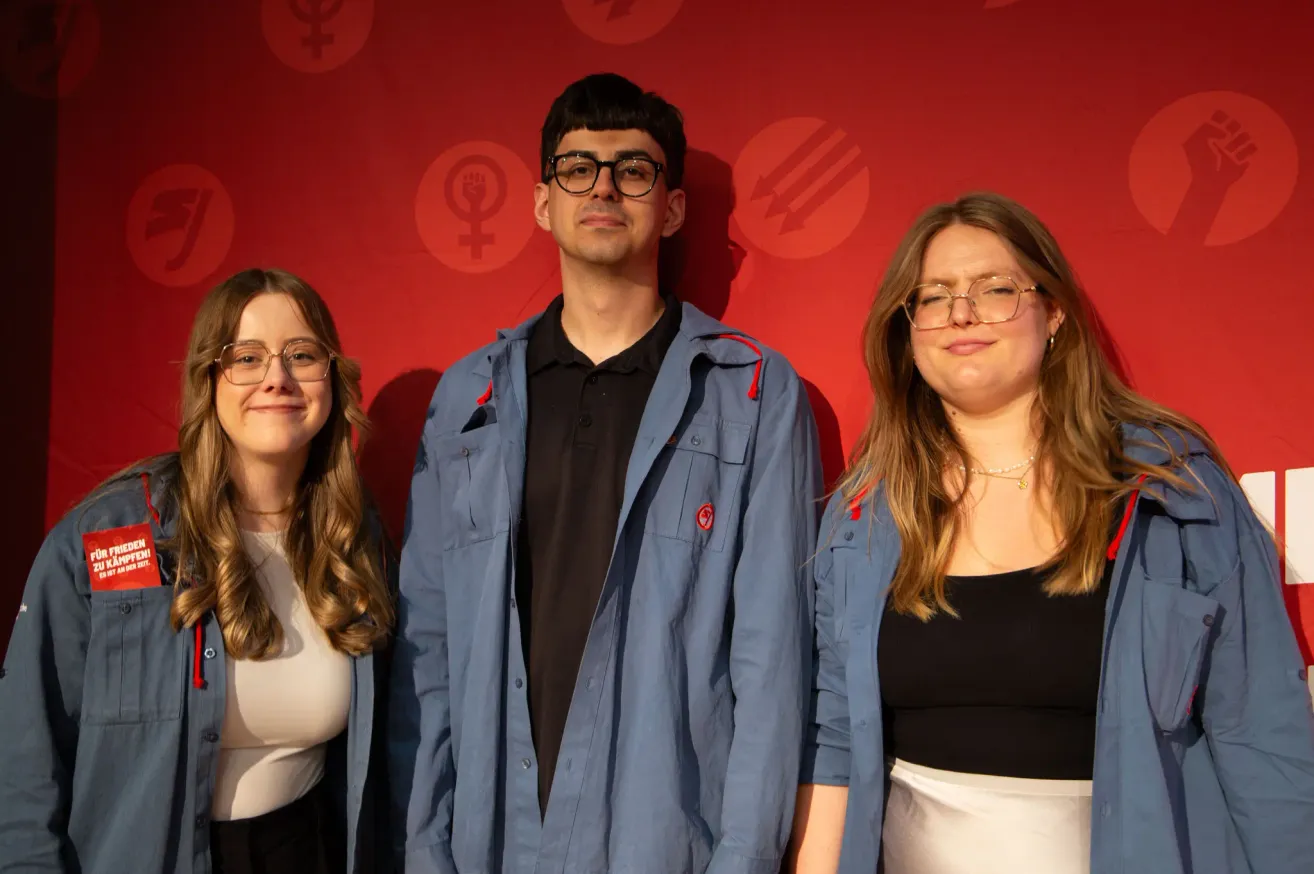 Three people, two women and a man, standing together in front of a red background with a symbol. They are all wearing glasses and are smiling.