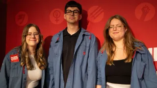 Three people, two women and a man, standing together in front of a red background with a symbol. They are all wearing glasses and are smiling.