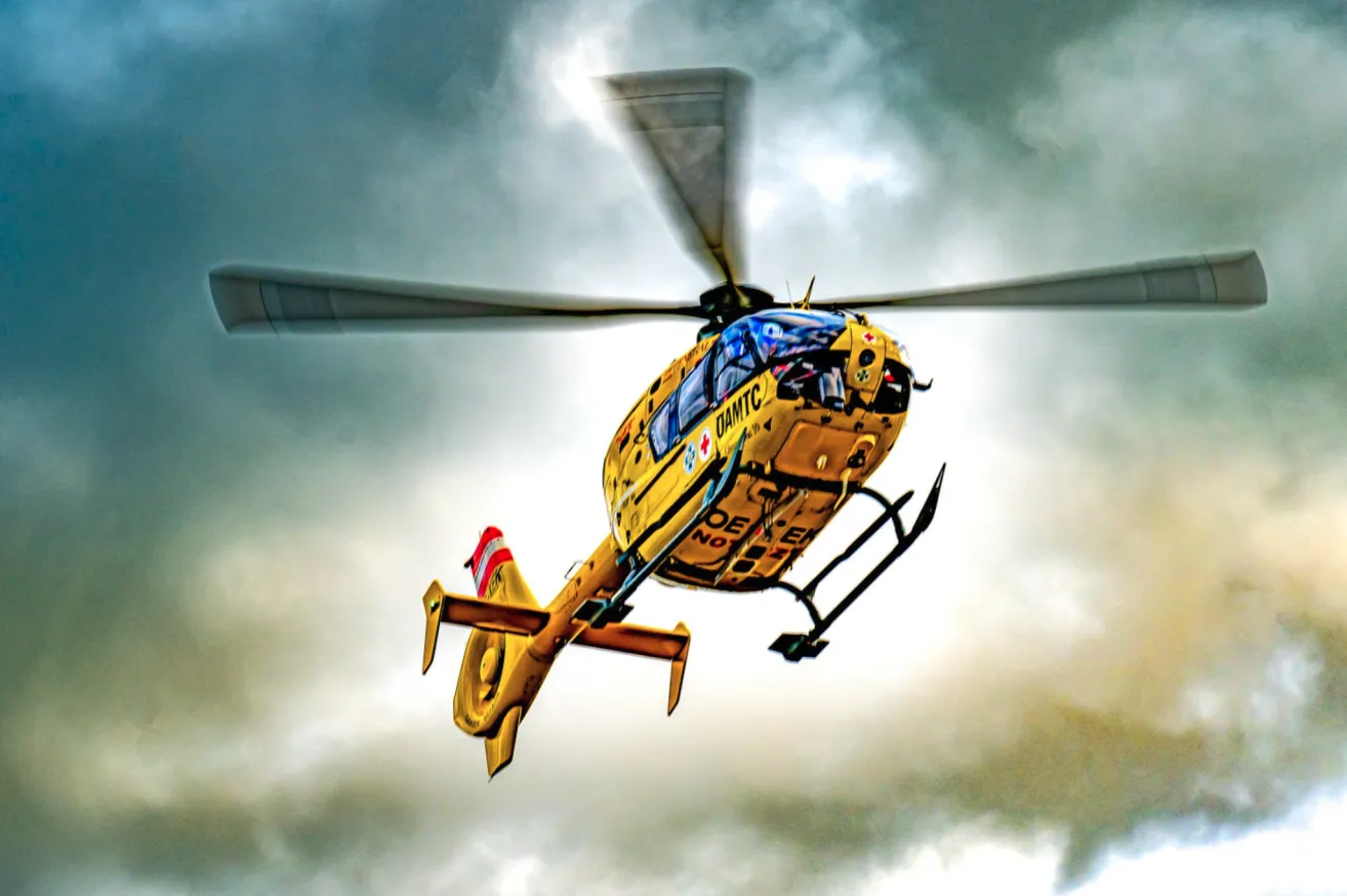 Ein gelber Hubschrauber mit der Aufschrift 'DAMTC' fliegt durch den bewölkten Himmel, mit einem roten und weißen Heck.