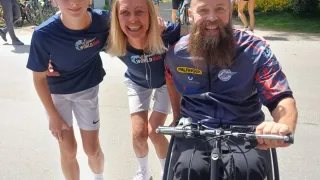 Three people pose for a photo. Two women wear World Run jerseys, and a man sits in a wheelchair. They are on a road with trees and people in the background.