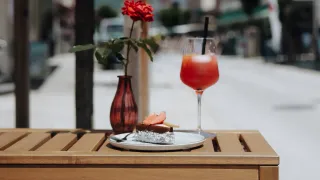 A dessert on a plate with a red rose in a vase and a glass of orange juice on a table outside.