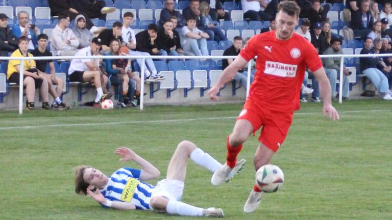 A soccer player in red chases the ball while another player in blue and white lies on the ground. Spectators watch from the bleachers.