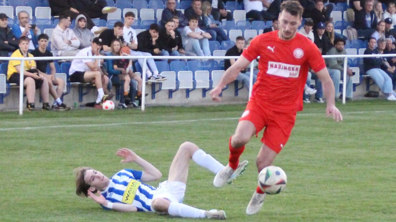 A soccer player in red chases the ball while another player in blue and white lies on the ground. Spectators watch from the bleachers.