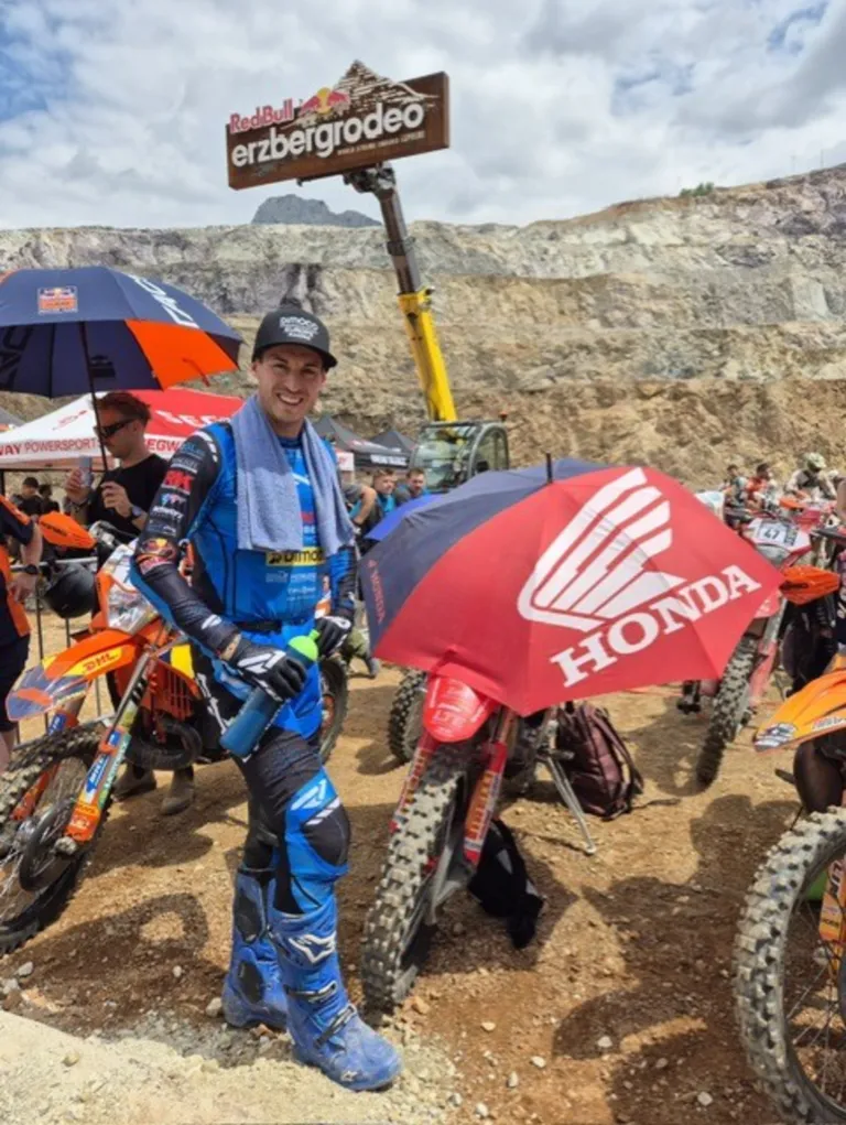 A man in blue gear stands with a Honda umbrella. He is surrounded by motorcycles and people on a dirt track.