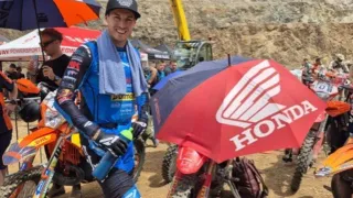 A man in blue gear stands with a Honda umbrella. He is surrounded by motorcycles and people on a dirt track.