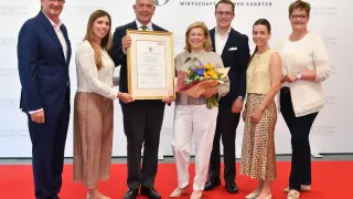 Eine Gruppe von Menschen, wahrscheinlich eine Familie, steht auf einem roten Teppich und posiert für ein Foto. Eine Frau in der Mitte hält einen Blumenstrauß und ein Zertifikat. Hinter ihnen befindet sich eine weiße Wand mit dem Text 'WKO' und '175'.