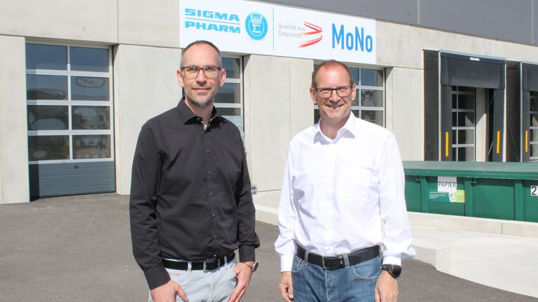 Zwei Männer stehen vor einem Gebäude mit einem Sigma Pharm Schild und einem MoNo Banner. Beide lächeln und sind formell gekleidet.