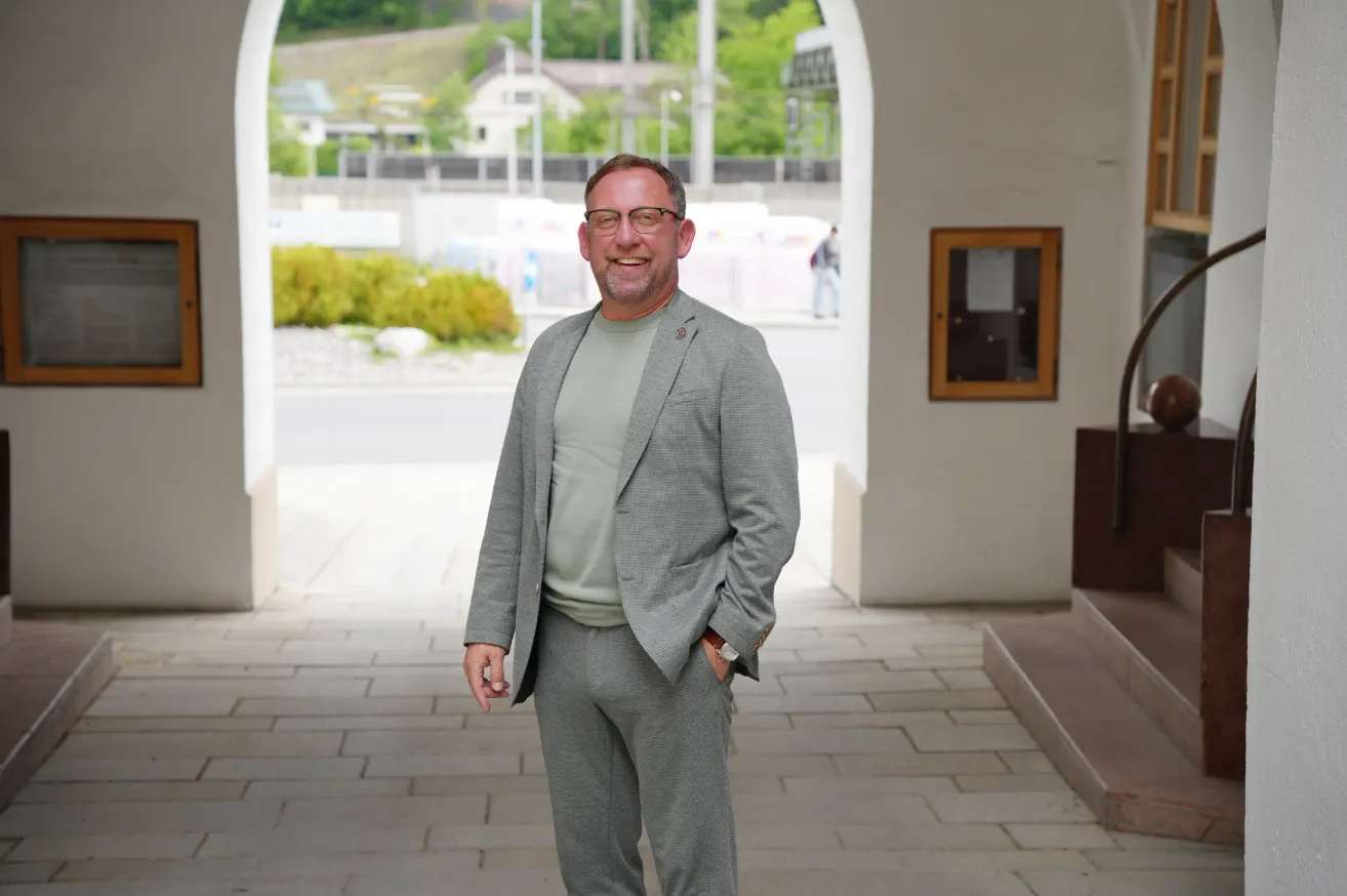 A man in a gray suit and glasses is smiling and standing in an archway. He has one hand in his pocket.