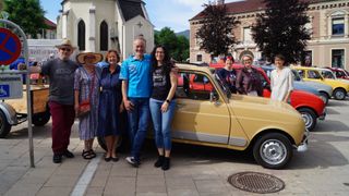 A group of people stands beside an old yellow car. They are dressed casually and are posing for a photo. Behind them, there are more cars, a church, and a building.