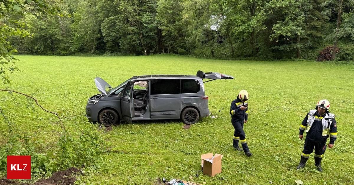 A damaged gray van with its hood open lies in a grassy field. A firefighter walks nearby, holding a radio.