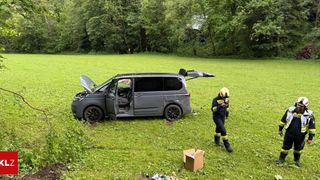 A damaged gray van with its hood open lies in a grassy field. A firefighter walks nearby, holding a radio.