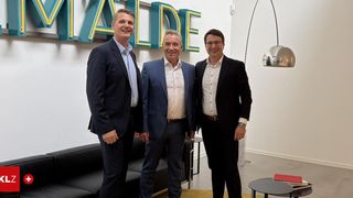 Three men in suits standing together in an office, smiling at the camera. Behind them, a white wall with the word 'WALDE' in large letters.