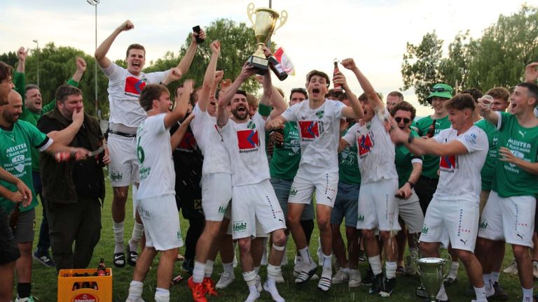 A soccer team celebrates on a grassy field, holding a trophy and bottles. Players wear matching white uniforms and some green ones.