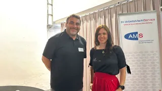 Two people stand smiling in front of a banner. The man wears a black polo, and the woman wears a black top and a red skirt. Behind them, a white curtain is draped.
