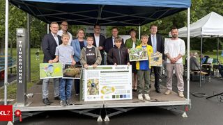 A group of adults and children stand under a tent holding posters and papers, some showing pictures of birds and insects. They are likely participating in a nature event.