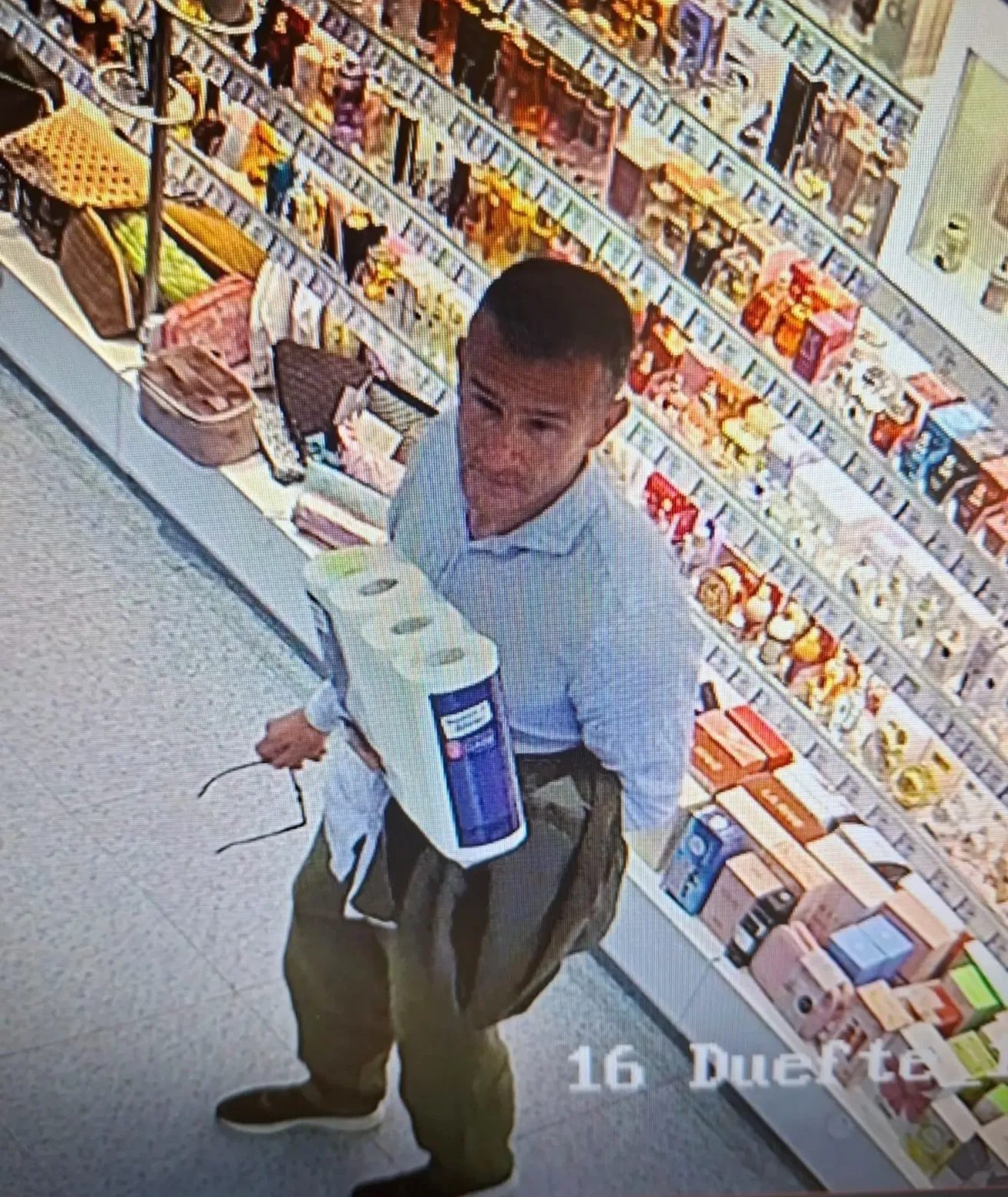 A man holds two toilet paper rolls in a store. He wears a blue shirt, glasses, and pants. Shelves behind him display various products.