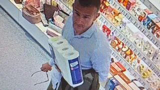 A man holds two toilet paper rolls in a store. He wears a blue shirt, glasses, and pants. Shelves behind him display various products.