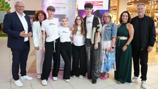 A group of people, likely students, wearing white t-shirts with text, stand in front of a banner. They are posing for a photo with an adult woman in a white coat.