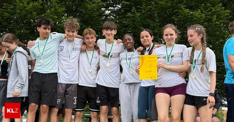 A group of young athletes in white shirts and medals poses for a photo. A girl holds a yellow certificate. Trees and a wooden bench are in the background.