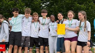 A group of young athletes in white shirts and medals poses for a photo. A girl holds a yellow certificate. Trees and a wooden bench are in the background.