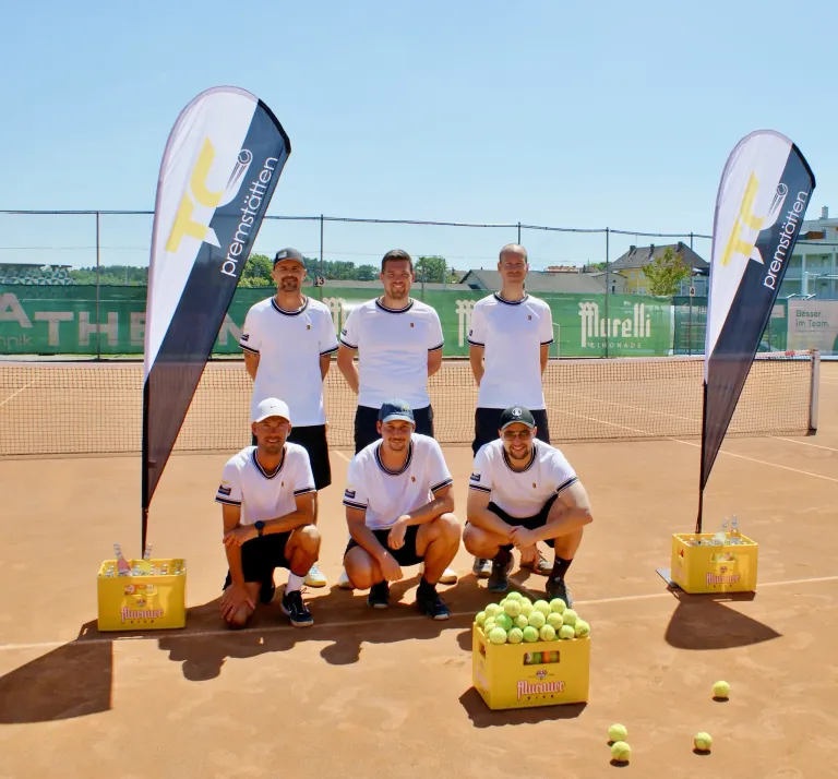 Sechs Männer in weißen Hemden und schwarzen Shorts posieren auf einem Tennisplatz mit zwei Bannern, Tennisbällen und Kisten. Der Platz ist von einem Zaun umgeben, mit Bäumen und Gebäuden in der Ferne.