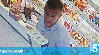 A man is seen in a supermarket, holding two rolls of tissue. He wears a light blue polo shirt. In the background, shelves filled with various products are visible.