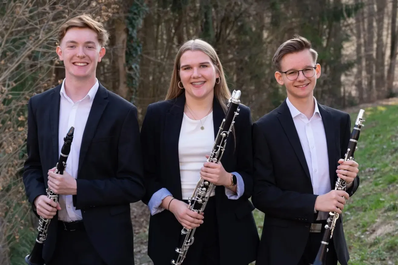 Three musicians dressed in black suits and white shirts pose for a picture, with one woman holding a clarinet.