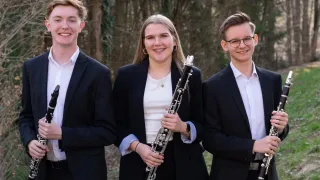 Three musicians dressed in black suits and white shirts pose for a picture, with one woman holding a clarinet.