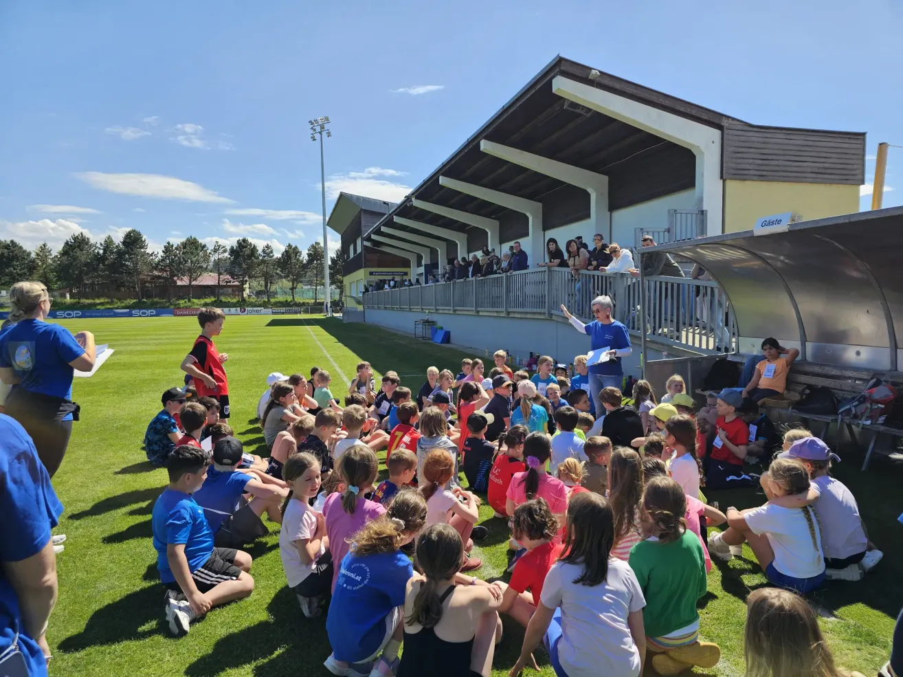 Eine Gruppe von Kindern sitzt auf dem Rasenfeld vor einem Stadion, wo ein Erwachsener zu ihnen spricht. Einige Leute stehen auf der Balkon des Stadions. Der Himmel ist blau mit einigen Wolken.