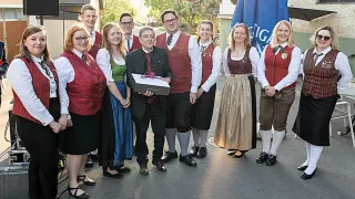 A group of people in traditional Bavarian attire pose for a photo, with a man holding a gift box. The setting appears to be outdoors.