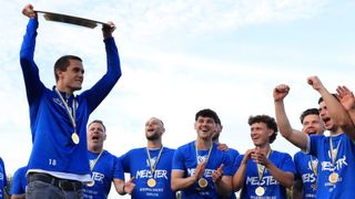 A group of men wearing blue shirts with gold medals celebrate with a silver trophy under a cloudy sky.